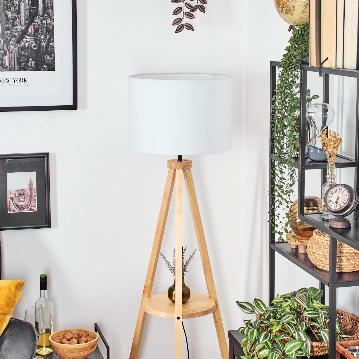 Eine Stehlampe mit weißem Lampenschirm und Holzbeinen. Im Hintergrund befindet sich ein Bücherregal mit Pflanzen und Dekoration.