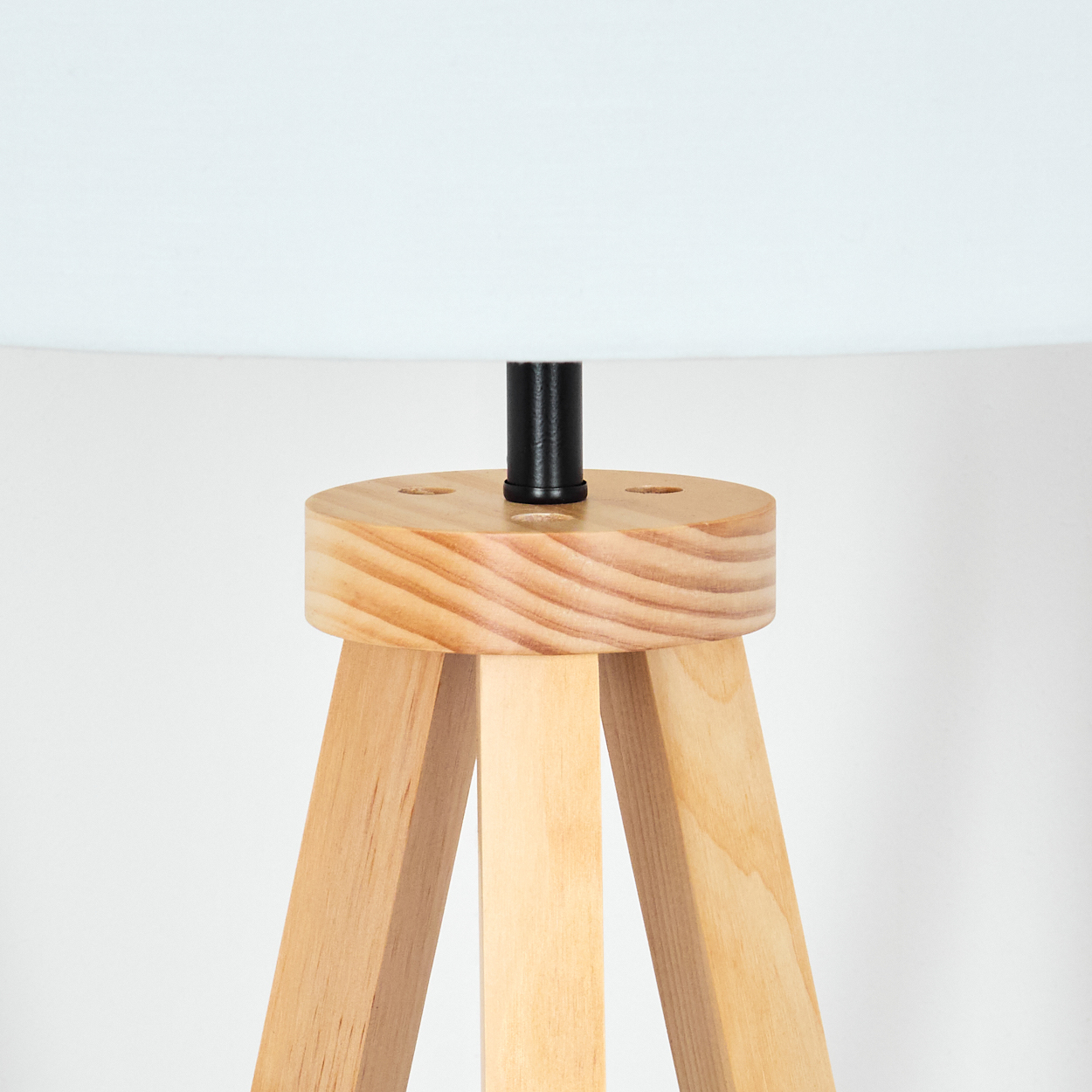 Eine hellfarbene Holzlampe mit weißem Lampenschirm. Der Fuß ist ein Dreibein. Die Lampe hat einen schwarzen Pfosten und einen hellen Holzsockel.