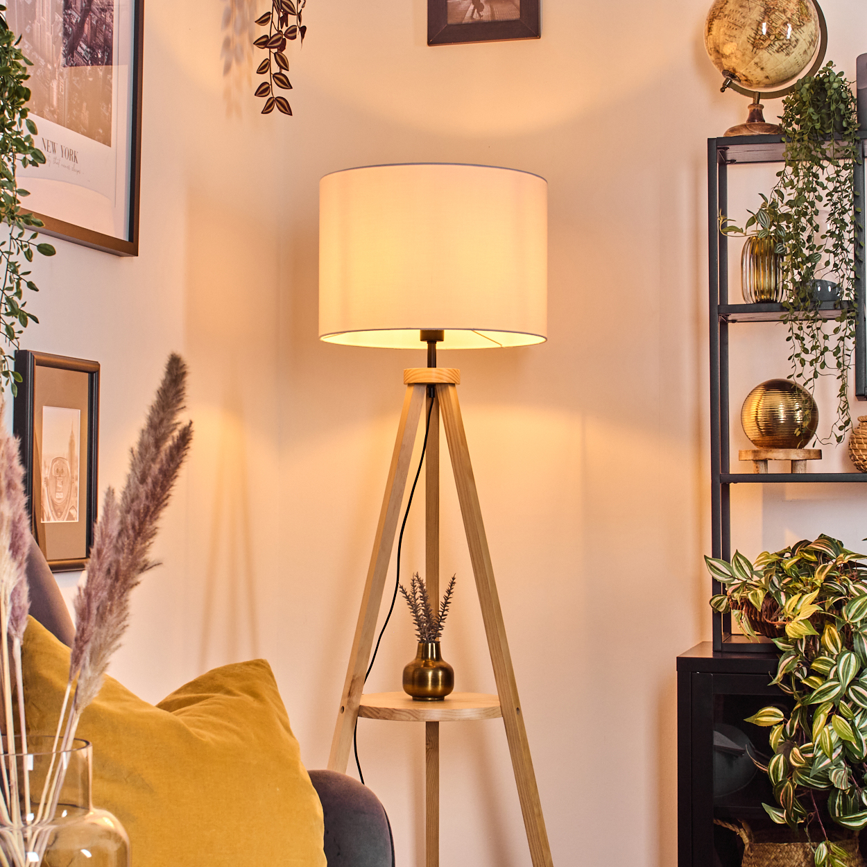 Eine beleuchtete hölzerne Stativlampe steht in der Nähe einer Couch mit gelbem Kissen und Pflanzen. Hintergrund: Globus, Regale mit Pflanzen und Dekor.