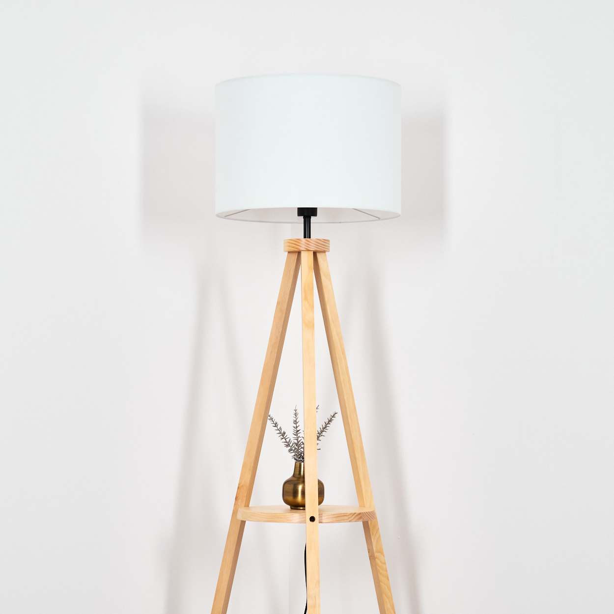 Eine Stehlampe aus Holz mit weißem Lampenschirm. Eine kleine goldene Vase mit getrockneten Pflanzen steht auf dem Regal.