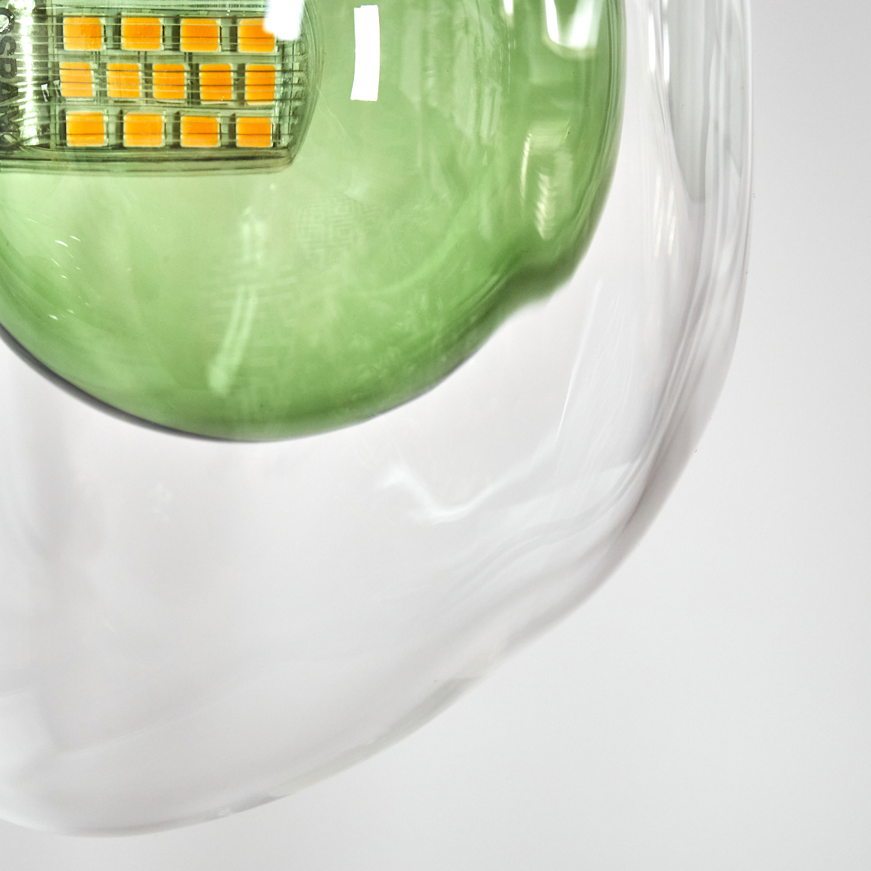 Nahaufnahme eines Glasobjekts. Eine grüne Kugel befindet sich in einem klaren Glas. Die Oberseite hat ein Quadrat mit kleinen orangen Lichtern. Weißer Hintergrund.