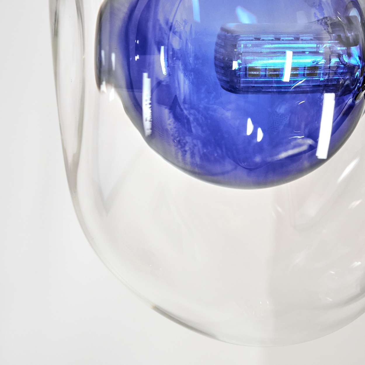 Ein klares Glasobjekt mit einem blauen, bauchigen Inneren. Der blaue Bereich reflektiert Licht, der Hintergrund ist weiß.