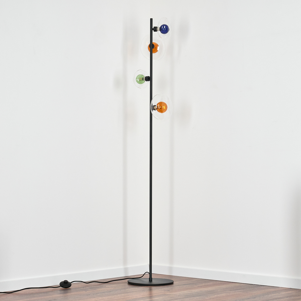 Eine schwarze Stehlampe mit vier bunten Glühbirnen steht in einer Ecke. Der Hintergrund sind weiße Wände und Holzboden.