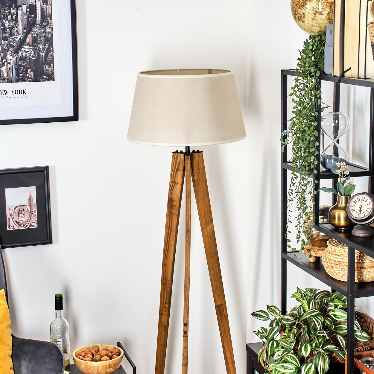 Eine Stehlampe mit beigem Lampenschirm steht neben einem schwarzen Regal mit Pflanzen, Büchern und Deko. Kunst hängt an der weißen Wand.