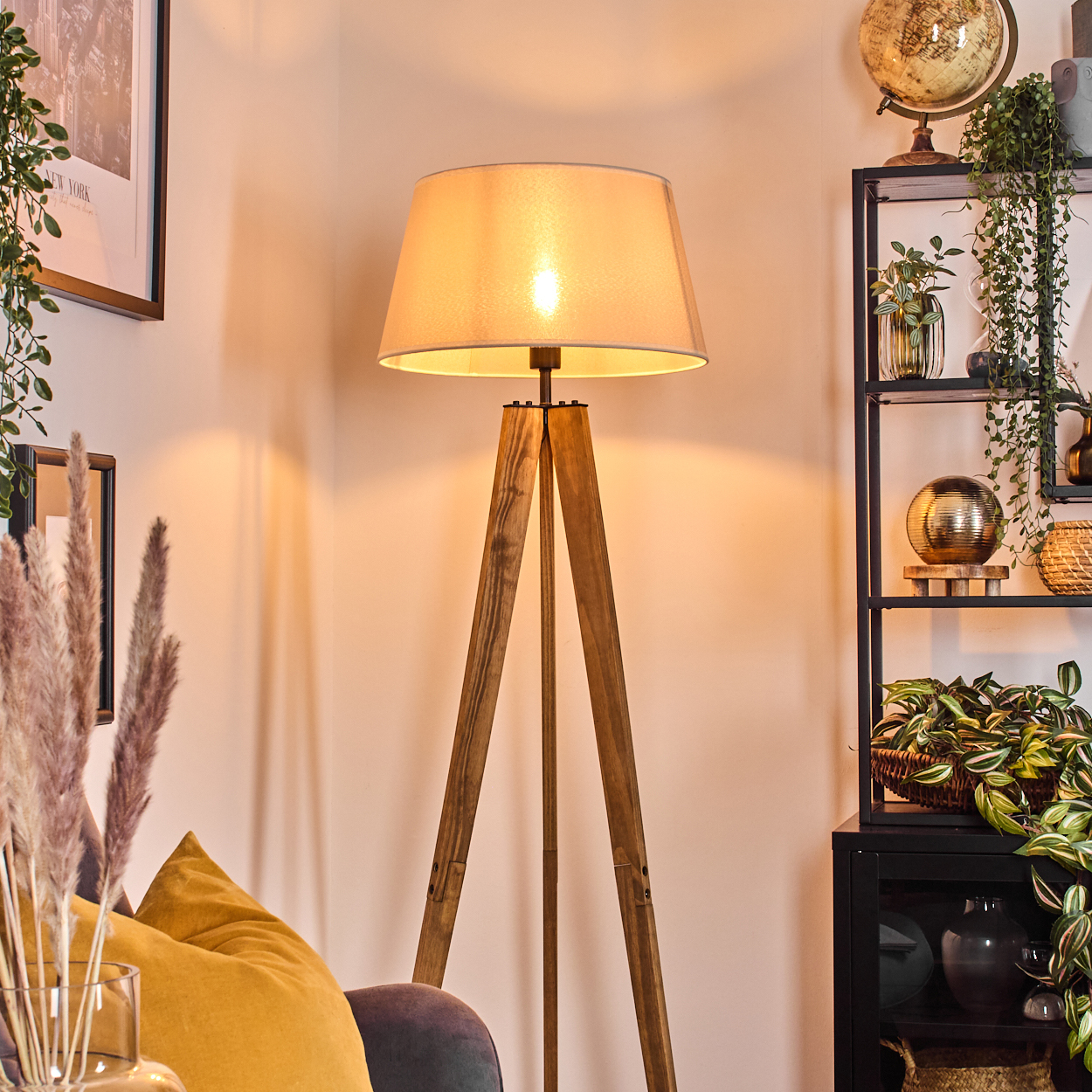 Eine Holzstativlampe leuchtet, mit Lampenschirm. Ein Regal mit Pflanzen und einem Globus. Ein gelbes Kissen und gerahmte Bilder.