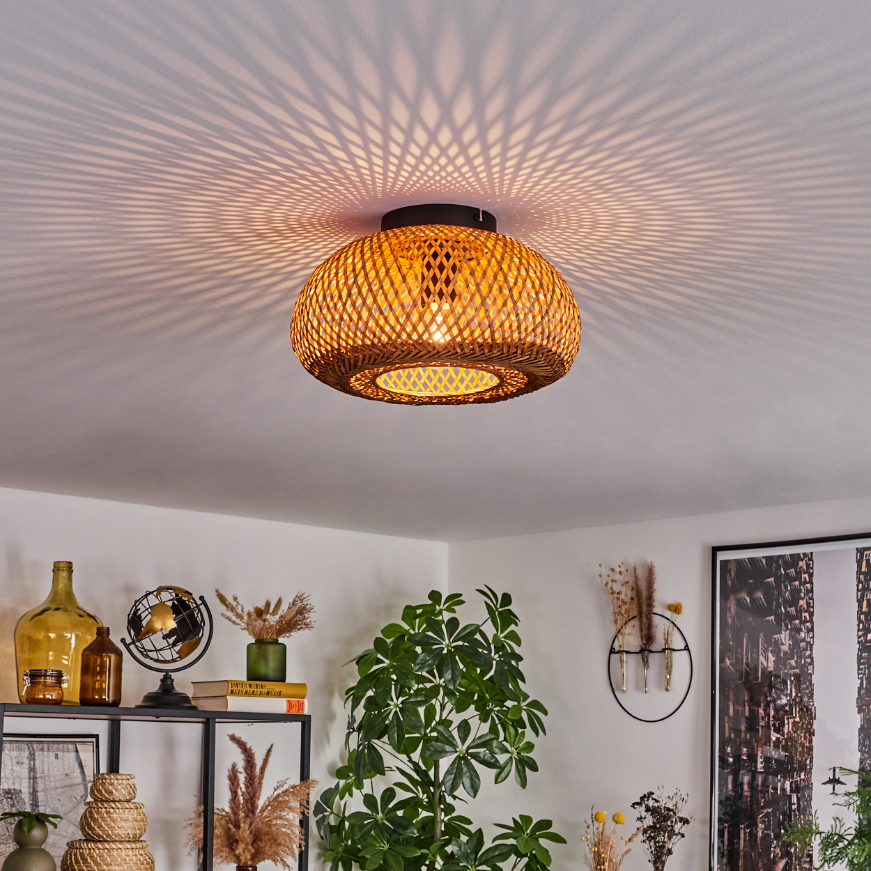 Eine gewebte Rattanlampe hängt an einer weißen Decke und wirft einen gemusterten Schatten. Darunter ein Regal mit Dekoration, Pflanzen und Kunst im Hintergrund.