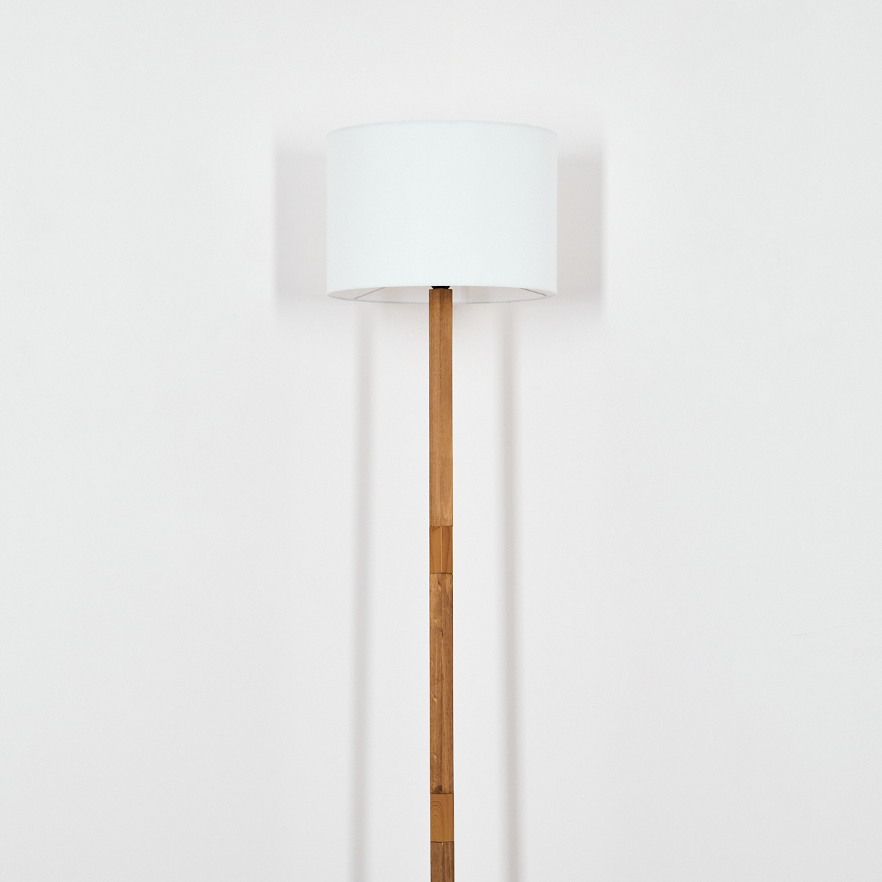 Eine hohe Lampe mit weißem Lampenschirm und Holzständer steht vor einer weißen Wand. Das Licht ist aus.