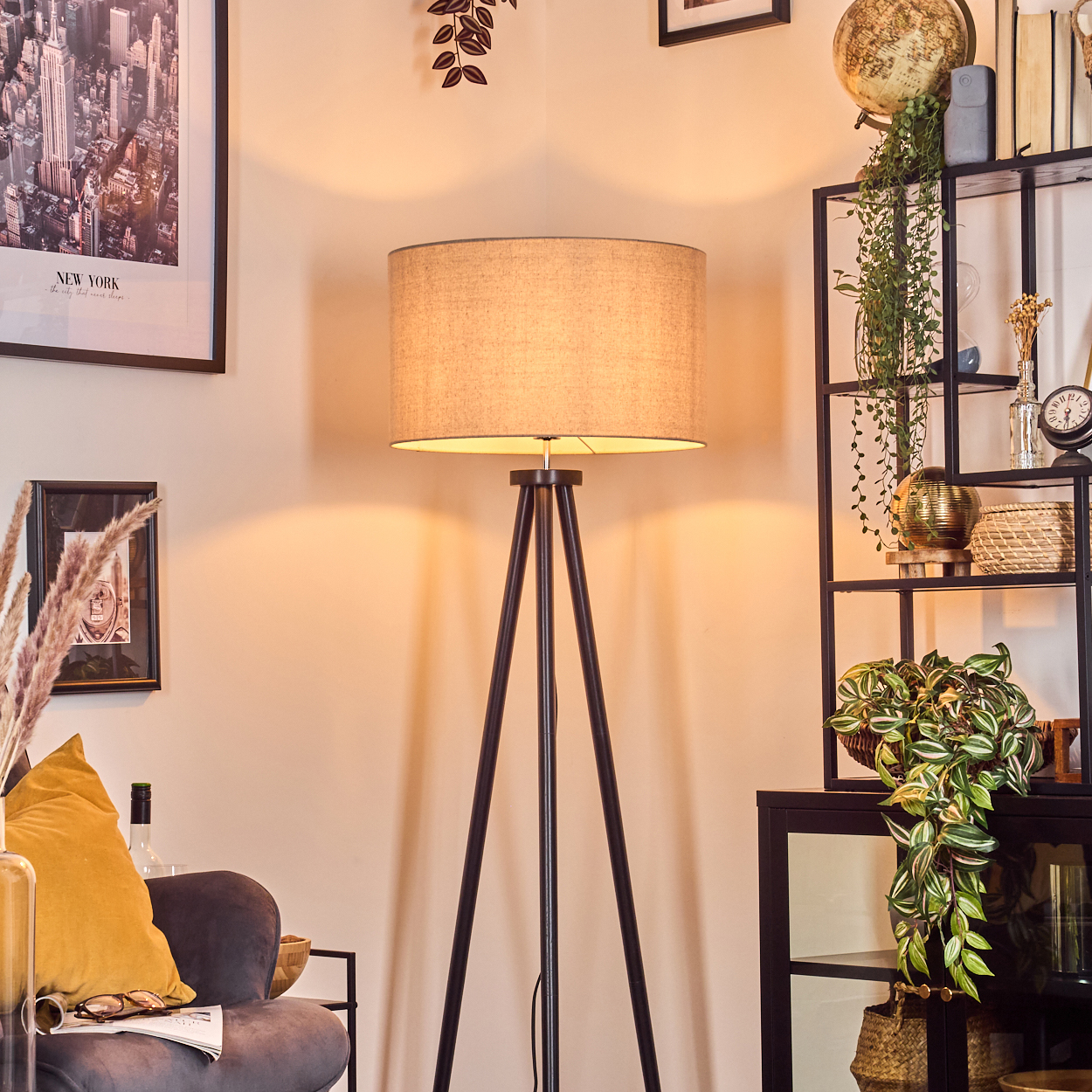 Eine Stehlampe mit beigem Lampenschirm steht in einem Raum. Rechts befindet sich ein schwarzes Regal mit Pflanzen und Dekoration.