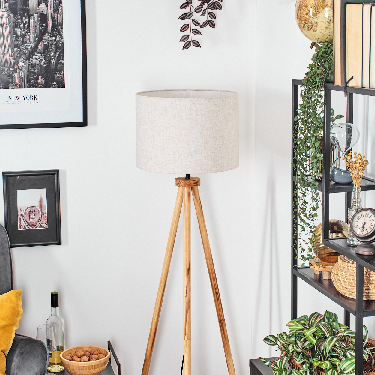 Eine Stehlampe mit beigem Schirm steht in einem Raum. Rechts ein schwarzes Regal mit Pflanzen, Büchern und einem Globus.