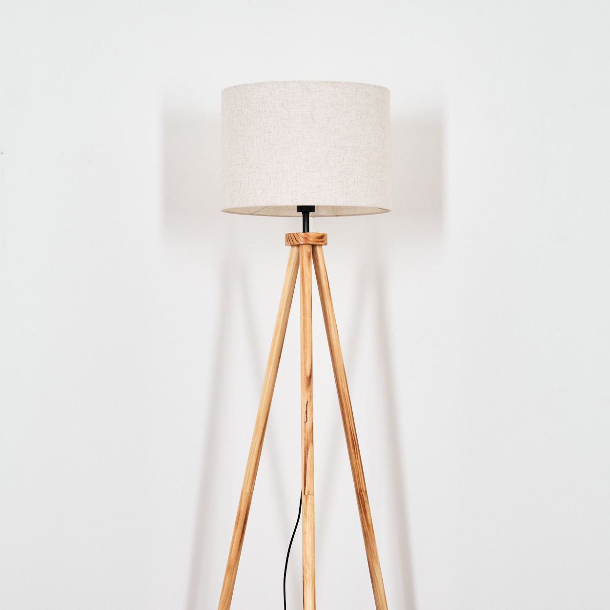 Eine Stehlampe aus Holz mit einem beigen Lampenschirm vor einer weißen Wand. Ein schwarzes Kabel verläuft am Bein.