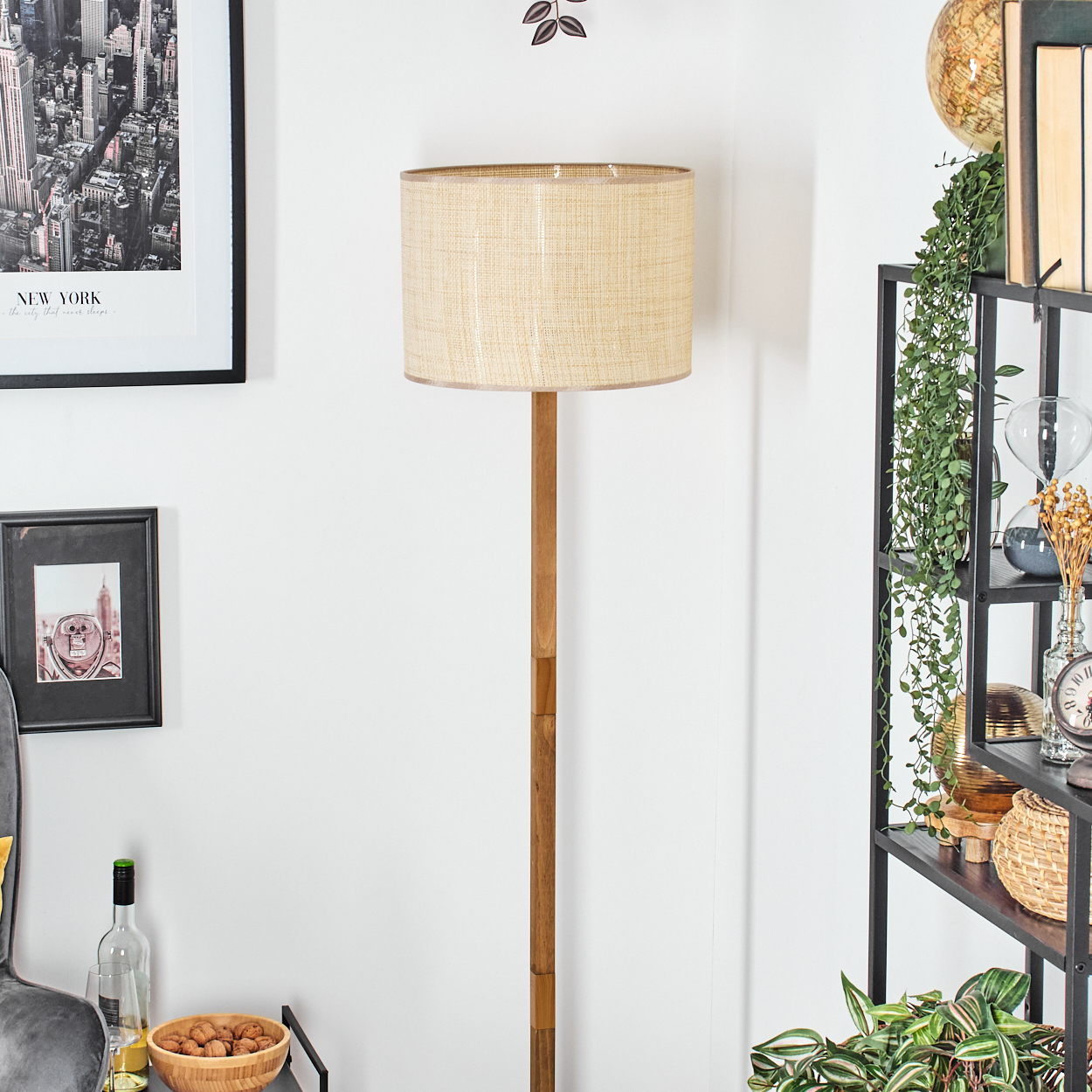 Eine Stehlampe mit beigem Schirm steht vor einer weißen Wand. Ein Regal mit Pflanzen und Dekorationen ist sichtbar.