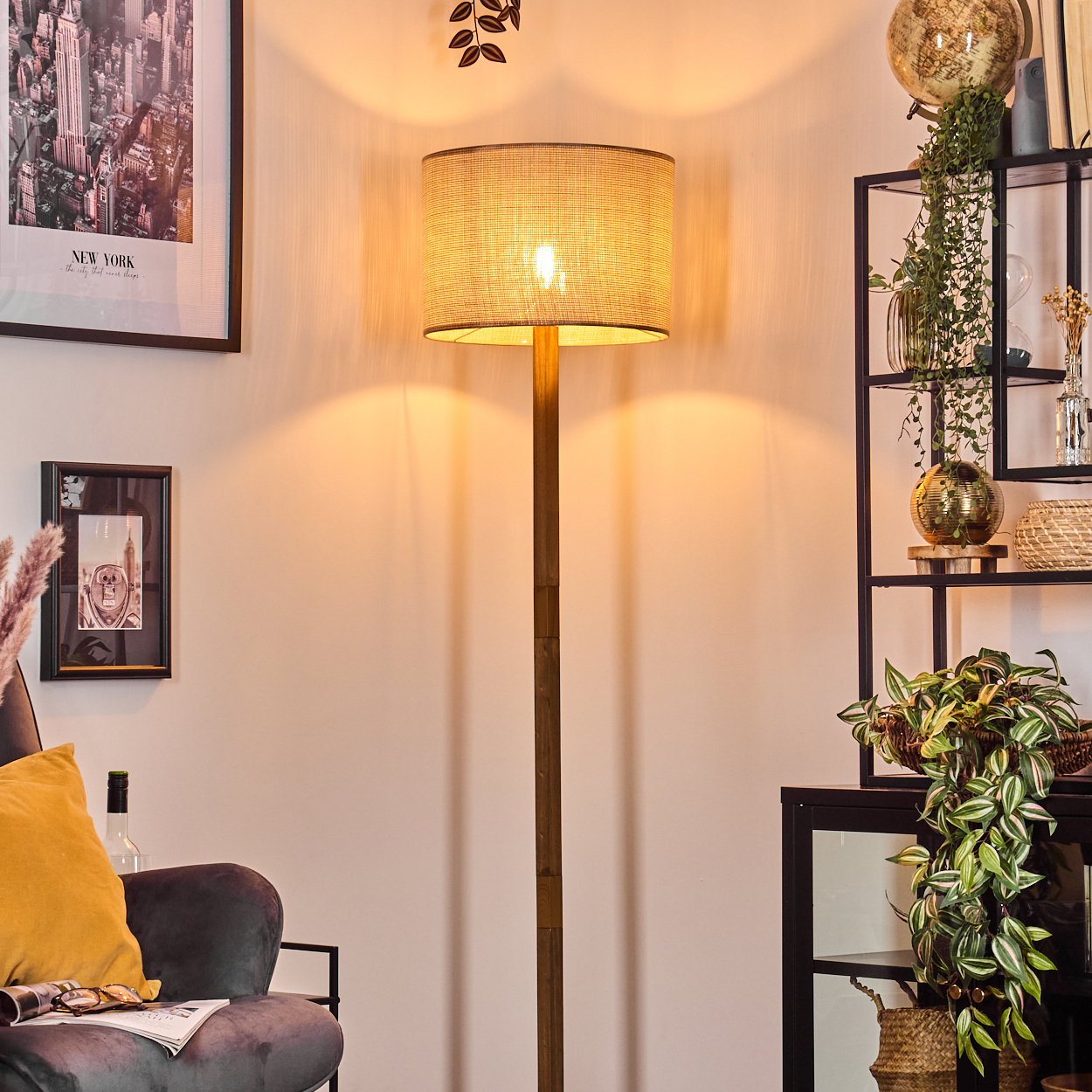 Eine beleuchtete Stehlampe steht in einem Raum mit Regal, Bildern und einer Couch. Die Lampe und das Regal stehen im Mittelpunkt.