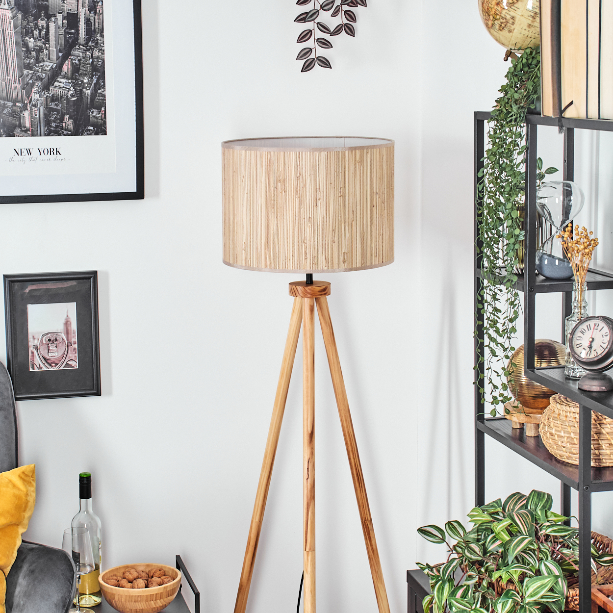 Eine hölzerne Stativ-Stehlampe steht in einem Raum. Rechts steht ein Regal mit Dekoration und Pflanzen. Ein Schwarz-Weiß-Foto von New York hängt an der Wand.