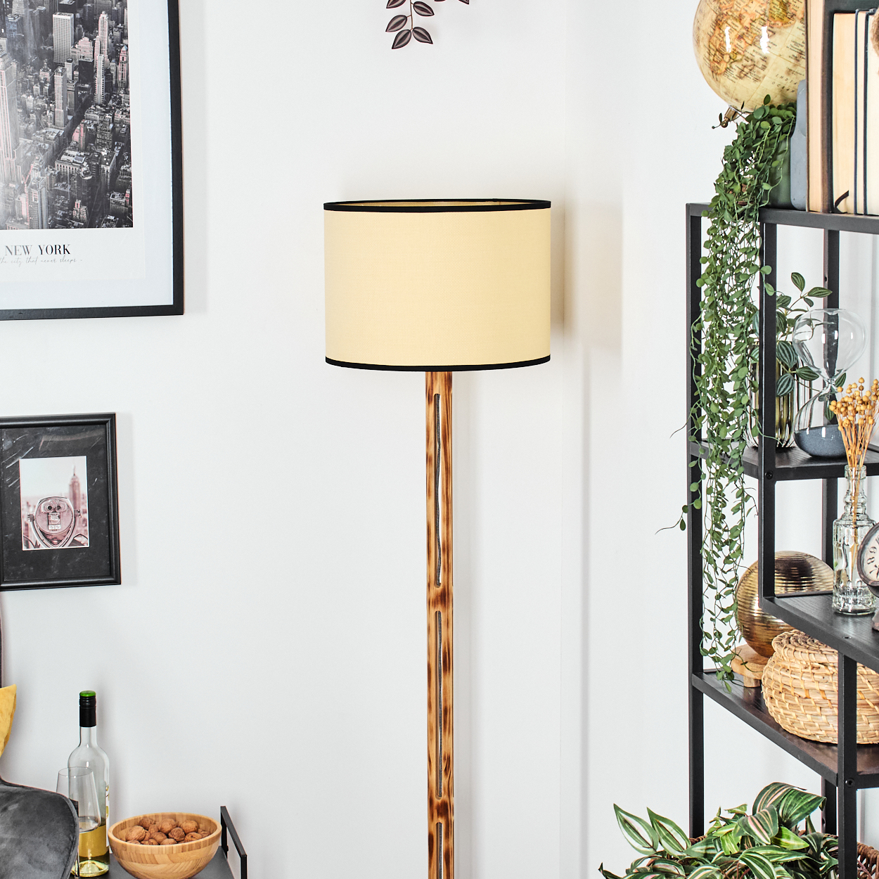 Eine Stehlampe mit beigem Lampenschirm und schwarzem Rand steht neben einem schwarzen Regal mit Pflanzen, Büchern und Dekorationsartikeln.