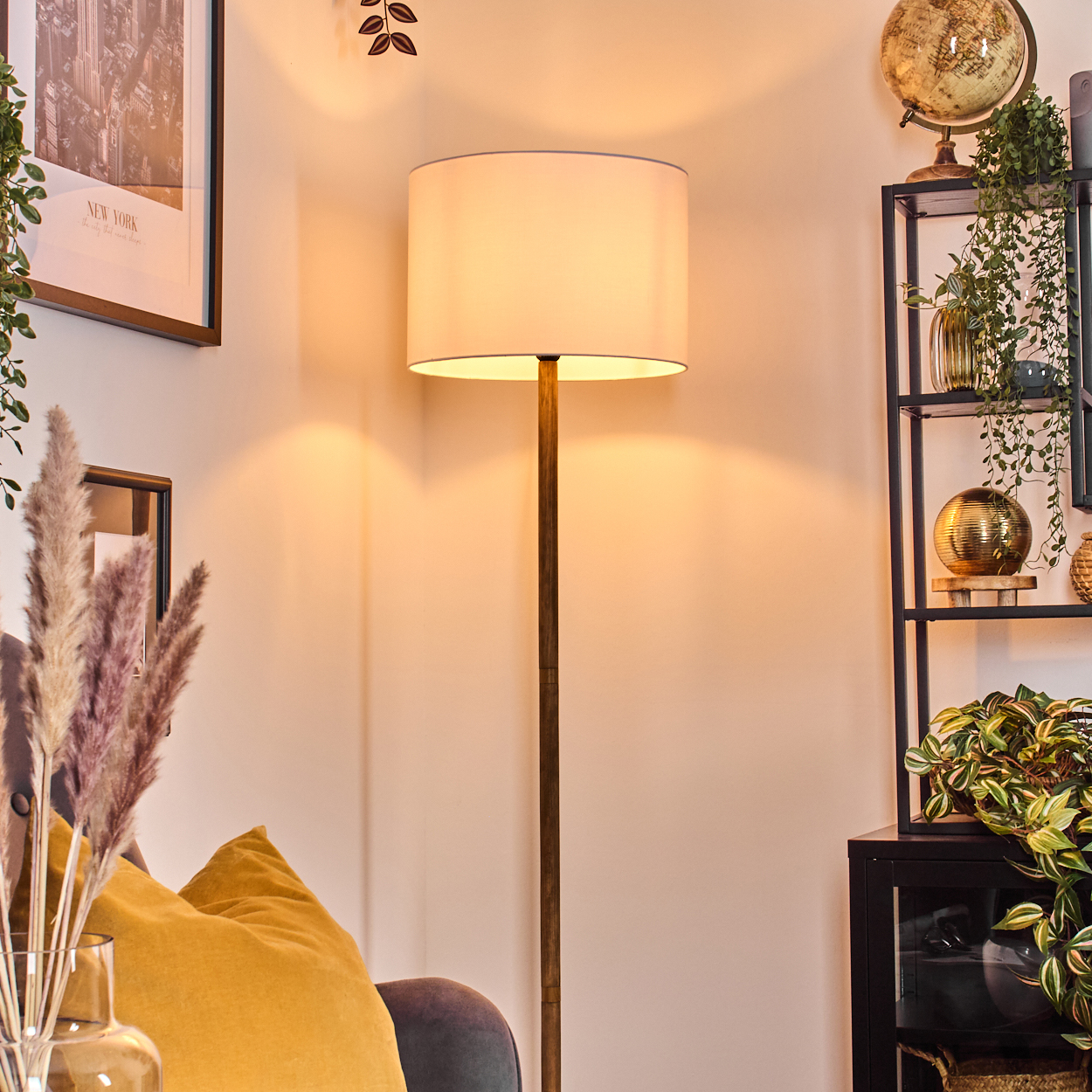 Eine beleuchtete Stehlampe steht neben einem Sofa mit gelben Kissen, neben einem Regal mit Pflanzen und Globus.