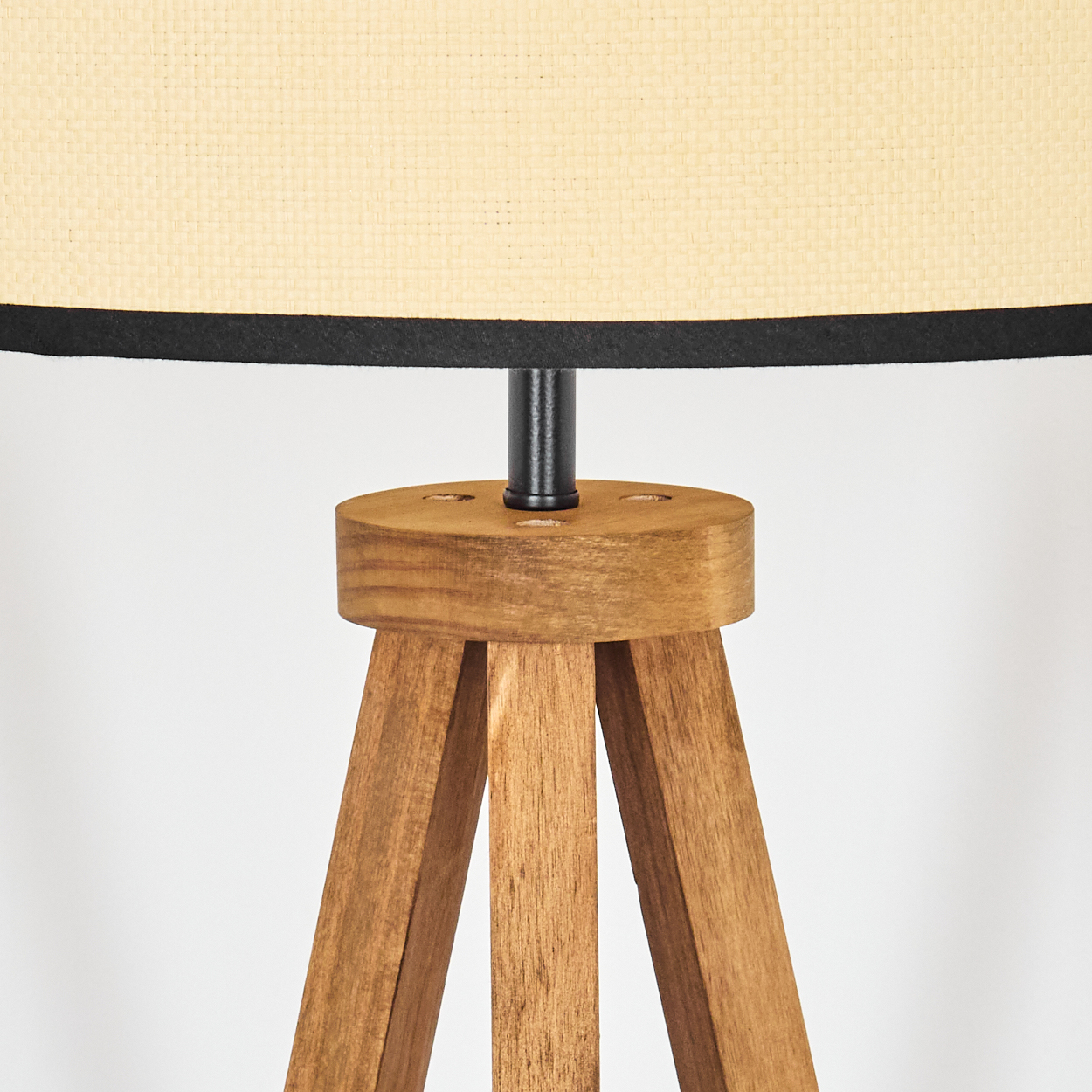 Eine Stehlampe mit Holzstativ, beigem Lampenschirm und schwarzem Rand. Die Lampe steht auf weißem Hintergrund. Die Holzbeine sind sichtbar.