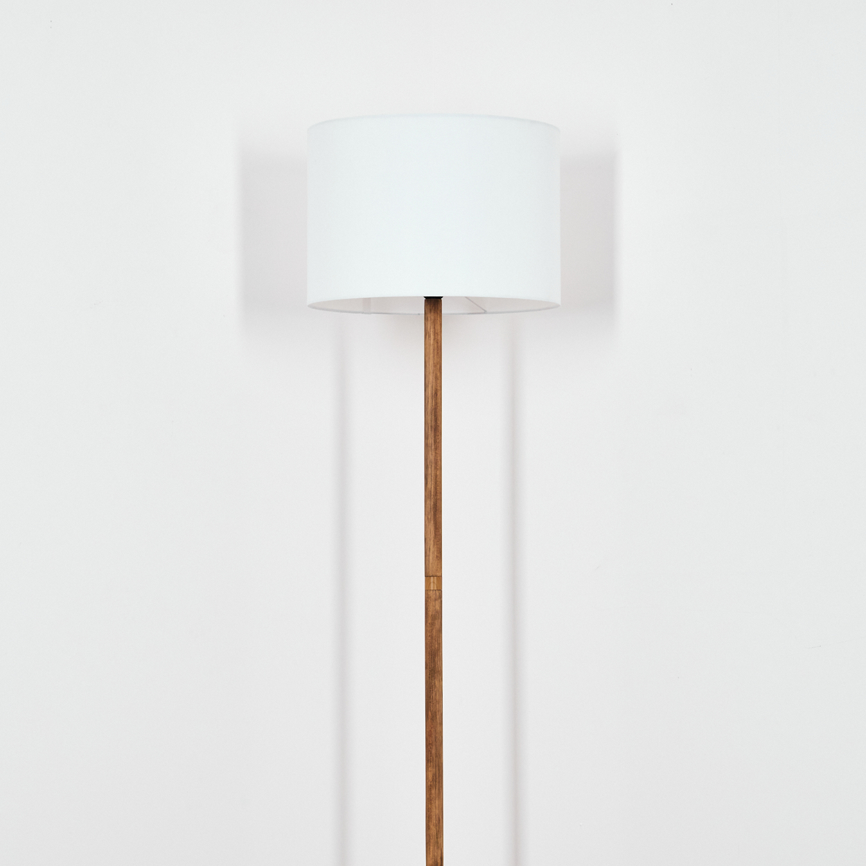 Eine Stehlampe mit weißem Lampenschirm und Holzstange steht vor einer weißen Wand. Die Lampe wirft einen Schatten.