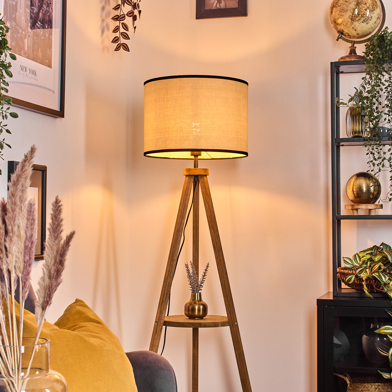 Eine hölzerne Stativ-Stehlampe mit beigem Schirm steht vor einer Wand. Ein Regal mit Pflanzen ist im Hintergrund.