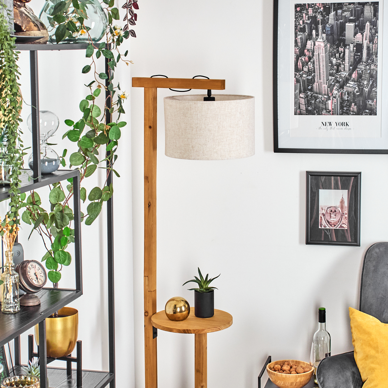 Zimmer mit Stehlampe, New York-Druck, Regalen mit Pflanzen und Dekor, einem grauen Sessel mit gelbem Kissen und einem Tisch.