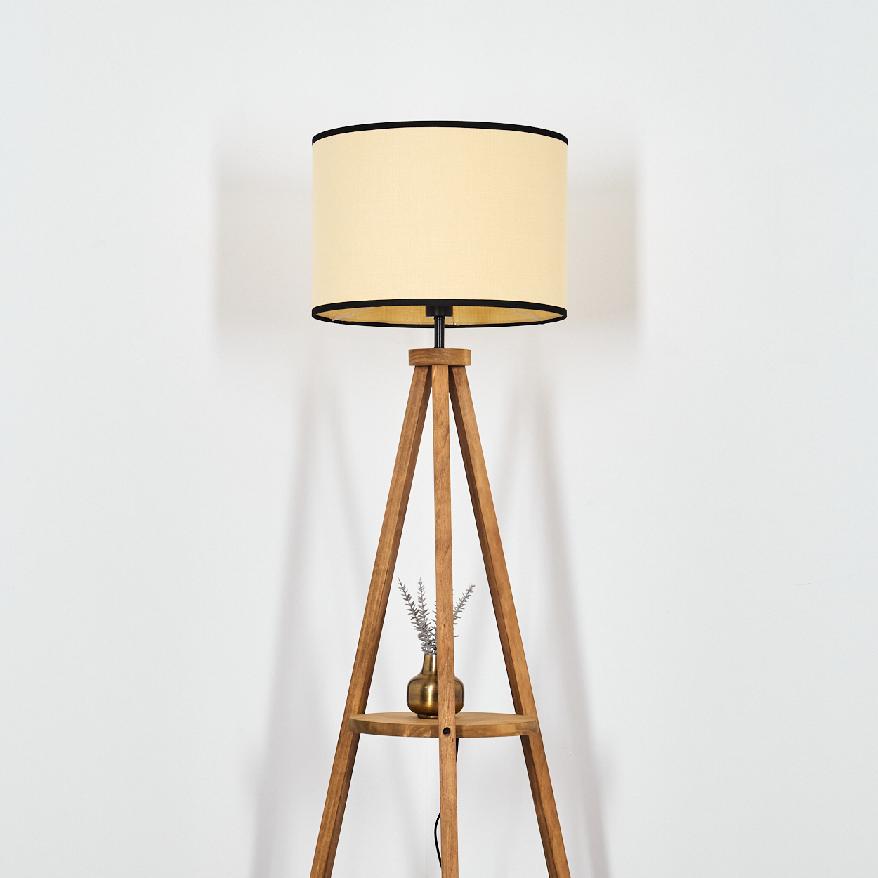 Eine Stehlampe aus Holz mit einem beigen Lampenschirm und schwarzem Rand. Eine kleine Vase mit Blumen steht auf einem Regal.