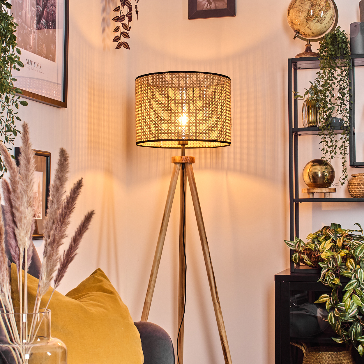 Eine beleuchtete gewebte Lampe steht auf Holzbeinen. Im Vordergrund befinden sich ein Sofa mit gelben Kissen und eine Vase mit Pampasgras.