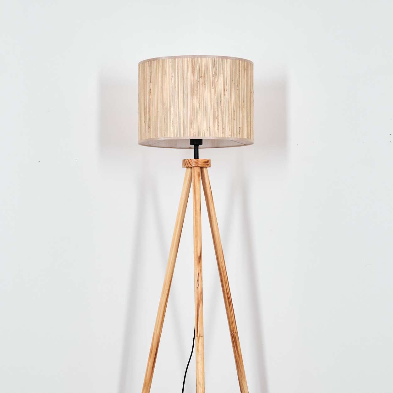Eine Stehlampe aus Holz mit Stativ und beigem, zylindrischem Lampenschirm steht vor einer weißen Wand.