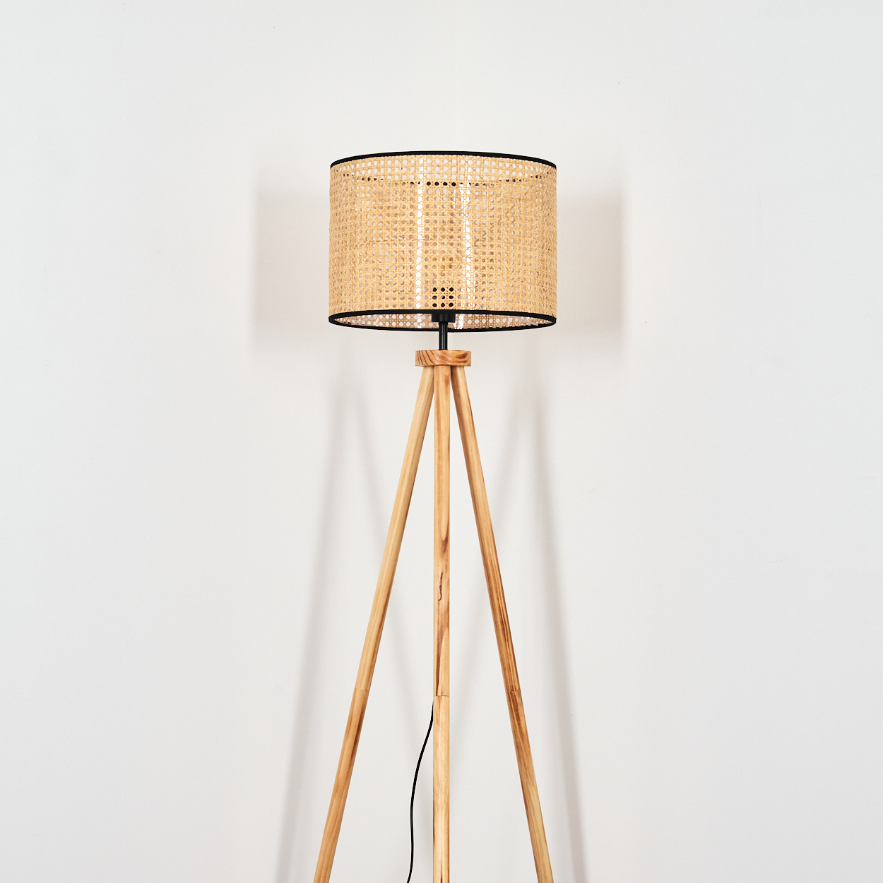 Eine hölzerne Stehlampe mit einem gewebten Lampenschirm steht vor einer weißen Wand. Ein schwarzes Kabel verläuft am Bein der Lampe.