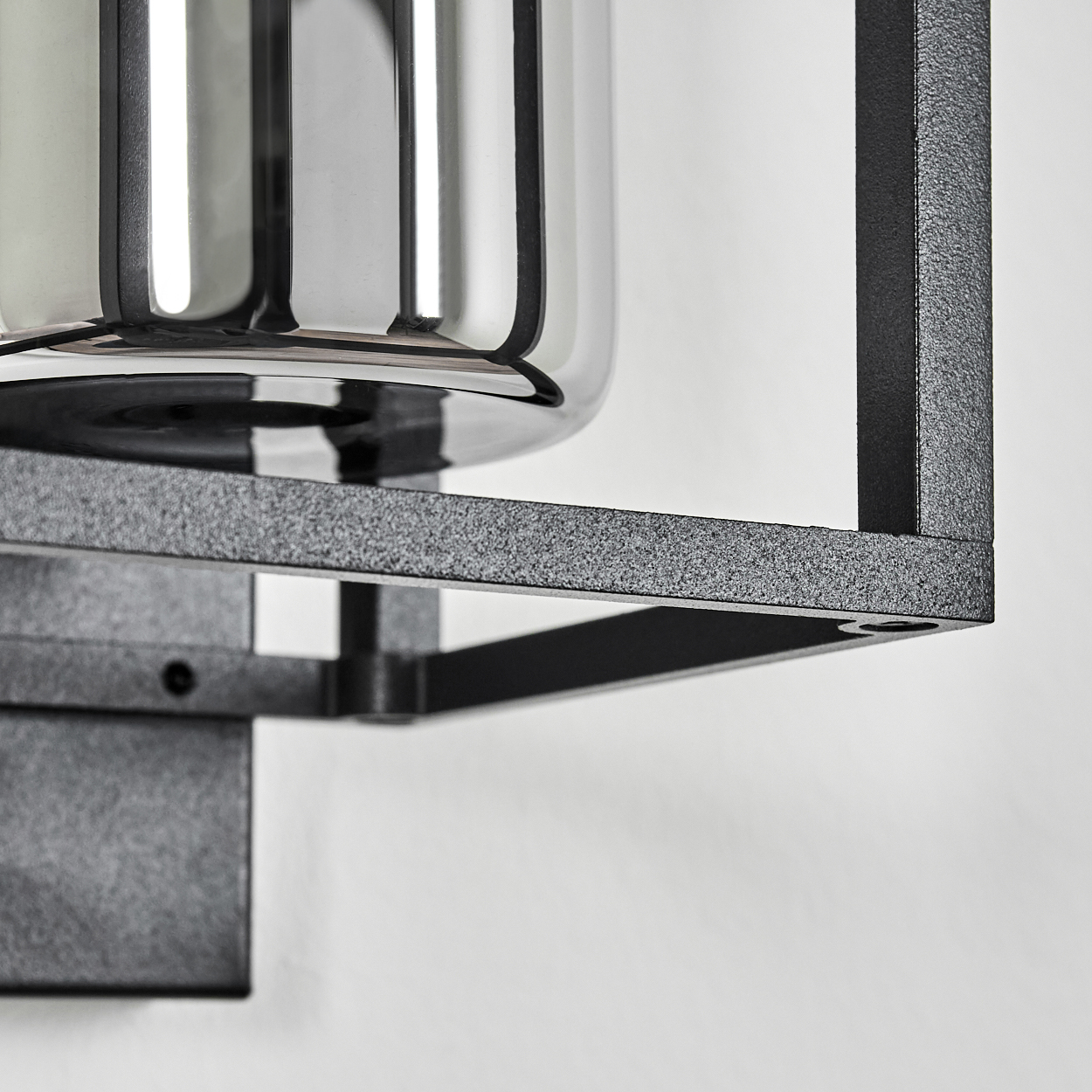 Nahaufnahme einer modernen Wandleuchte. Sie hat einen Metallrahmen mit einem Glaseinsatz. Der Rahmen ist dunkelgrau, der Hintergrund weiß.