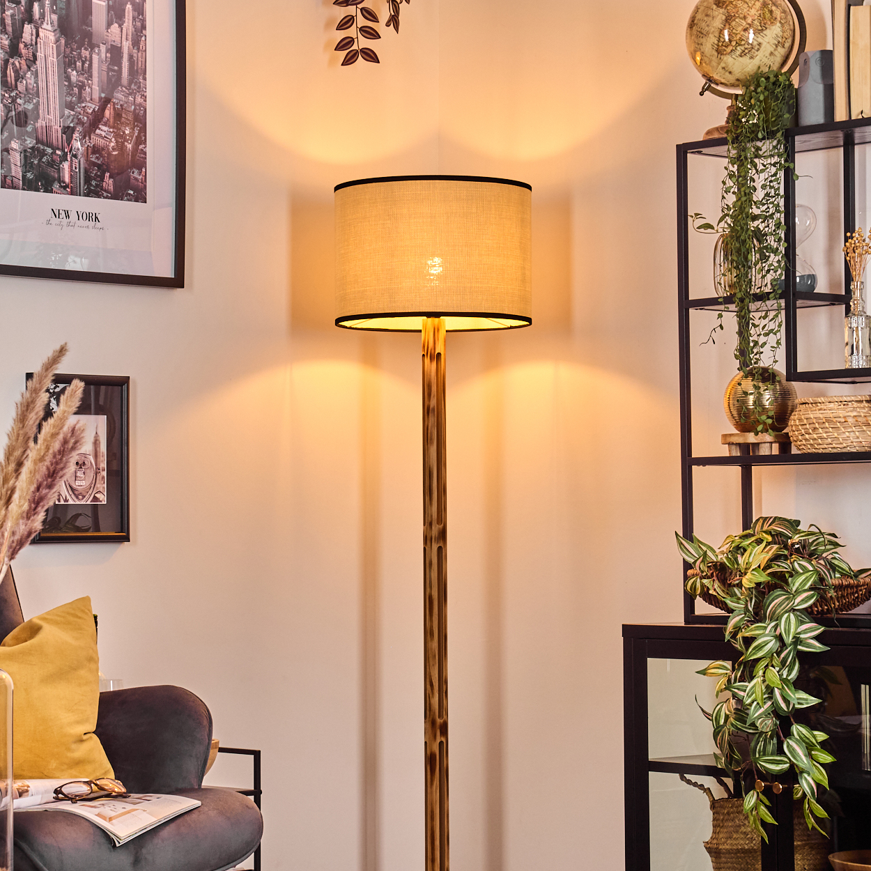 Eine beleuchtete goldene Stehlampe steht in einem Raum mit einem Poster von New York. Im Hintergrund befinden sich ein Globus und Regale mit Pflanzen.