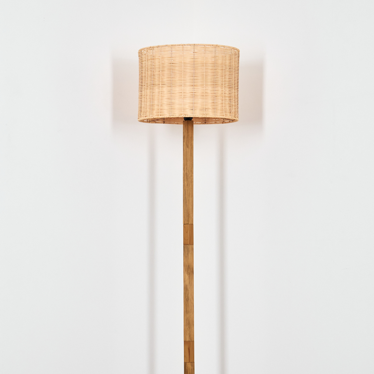 Eine Stehlampe mit gewebtem Lampenschirm und Holzstange vor einer weißen Wand. Die Lampe wirft einen Schatten.