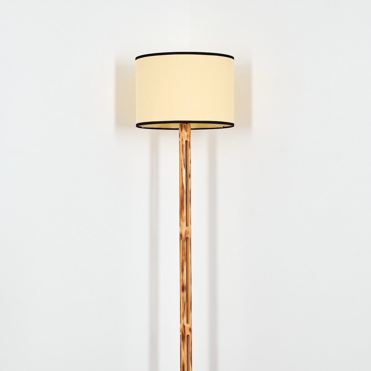 Eine Stehlampe mit beigem Lampenschirm, schwarzem Rand und einer braunen Bambusstange steht vor einer weißen Wand.