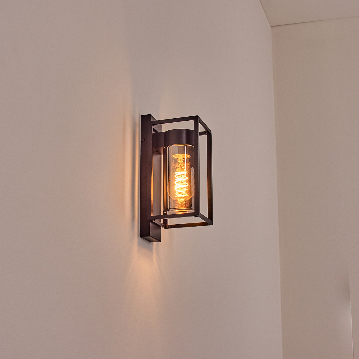 Eine schwarze Wandleuchte mit zylindrischem Glasschirm und einer leuchtenden Glühbirne vor weißer Wand.