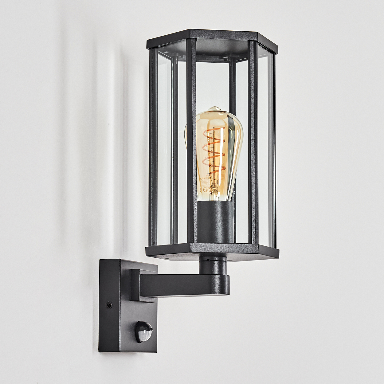 Schwarze Außenwandleuchte mit Glasscheiben, Edison-Glühbirne und Bewegungsmelder, an einer weißen Wand montiert.