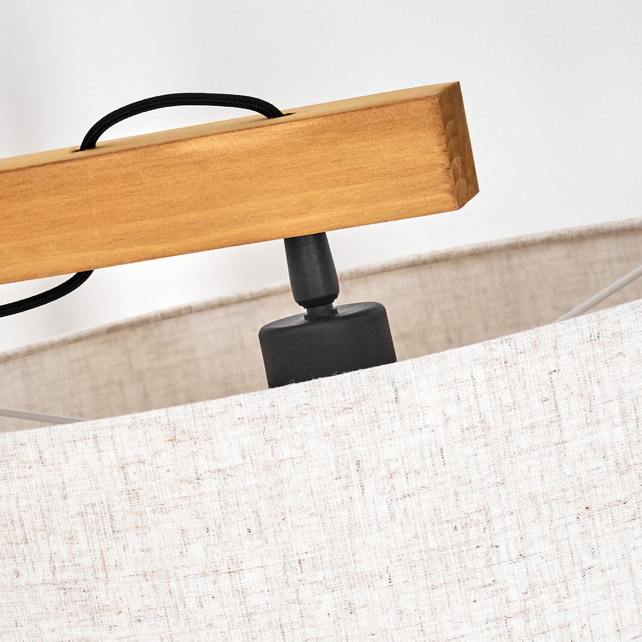 Nahaufnahme einer Lampe. Ein Holzbalken, ein schwarzes Kabel, eine schwarze Halterung und ein beiger Lampenschirm sind vor weißem Hintergrund sichtbar.
