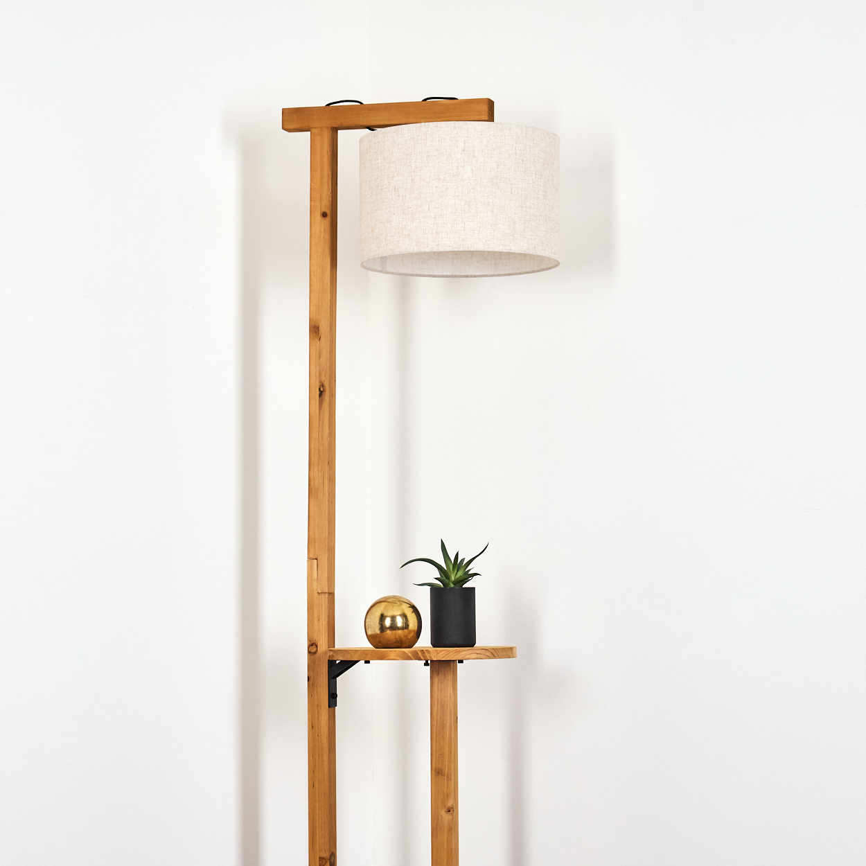 Eine hölzerne Stehlampe mit beigem Lampenschirm. Sie hat ein kleines Regal mit einer Pflanze und einer goldenen Kugel vor einer weißen Wand.