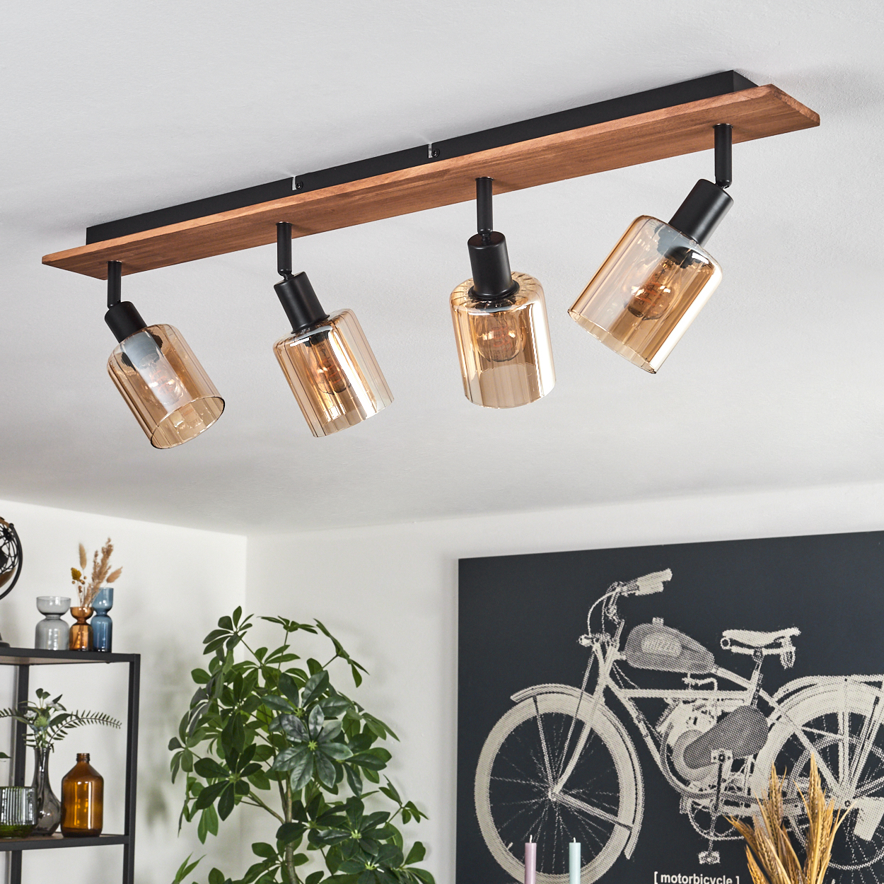 Eine Deckenleuchte mit vier bernsteinfarbenen Glasschirmen. Sie hängt an einer Holz- und Metallbefestigung. Eine Pflanze und Kunst sind im Hintergrund.