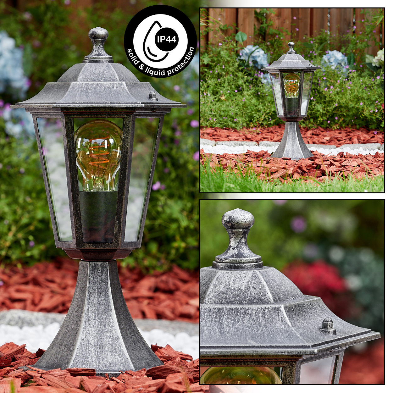 Eine silberne Außenlampe mit Glühbirne steht auf roter Mulch. Der Hintergrund zeigt Pflanzen und einen Holzzaun.