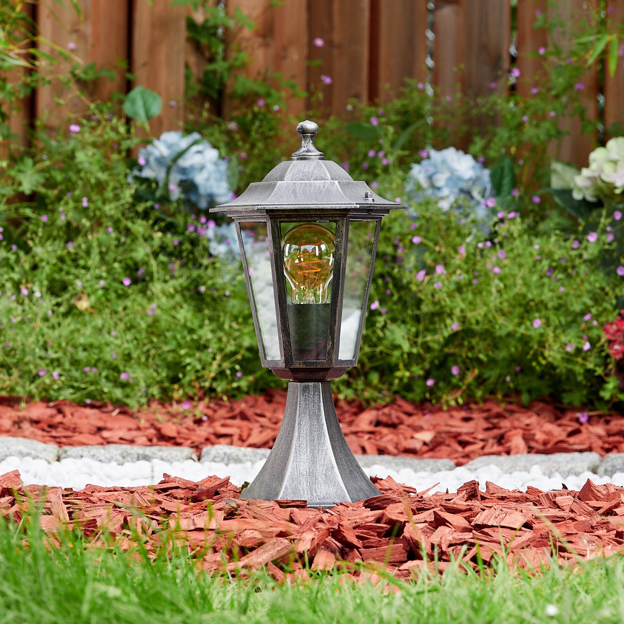 Eine silberne Außenlampe mit leuchtender Glühbirne steht in einem Gartenbeet mit roter Mulch, grünem Gras und Büschen.