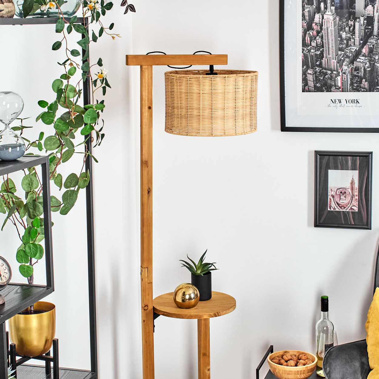 Eine hölzerne Stehlampe mit einem gewebten Lampenschirm steht neben einem Regal mit Pflanzen und Dekoration. Ein New Yorker Stadtbild hängt an der Wand.