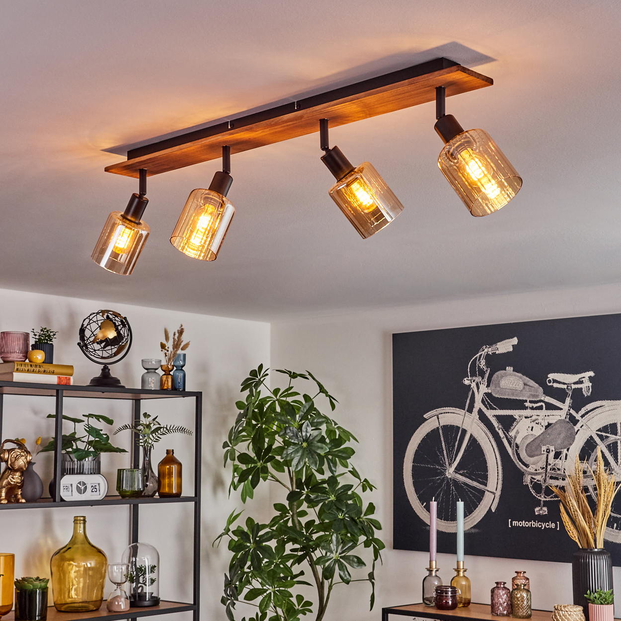Deckenleuchte mit vier Lampen. Sie ist an einem Holzbalken montiert. Der Raum hat Pflanzen, Regale mit Dekoration und ein Fahrradkunstwerk.