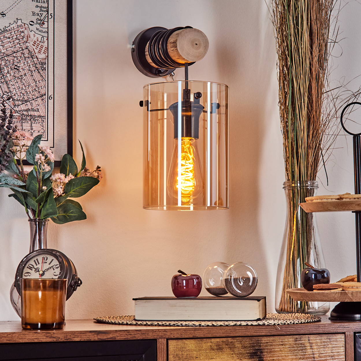 Eine leuchtende Edison-Glühbirnenlampe hängt. Darunter ein Tisch mit Deko: eine Kerze, ein Apfel, ein Buch und Vasen. An der Wand hängt eine Karte.