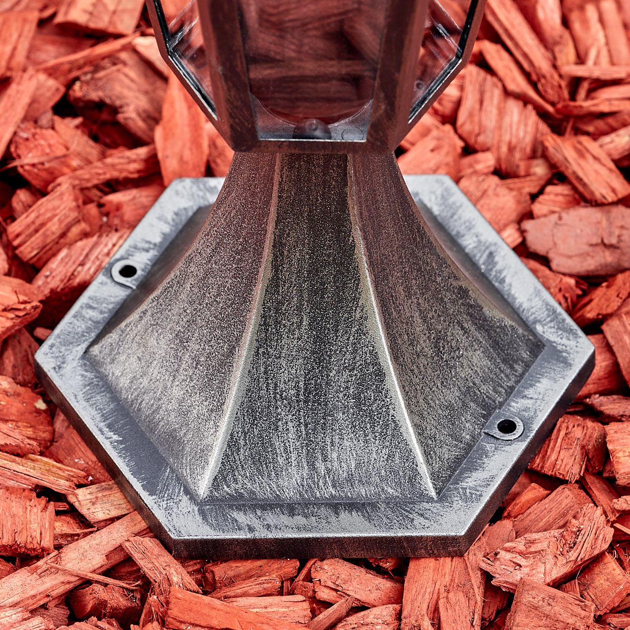 Nahaufnahme eines dekorativen Metallsockels auf roter Mulch. Der Sockel hat eine sechseckige Form mit Metallfinish. Die Mulch ist rot.