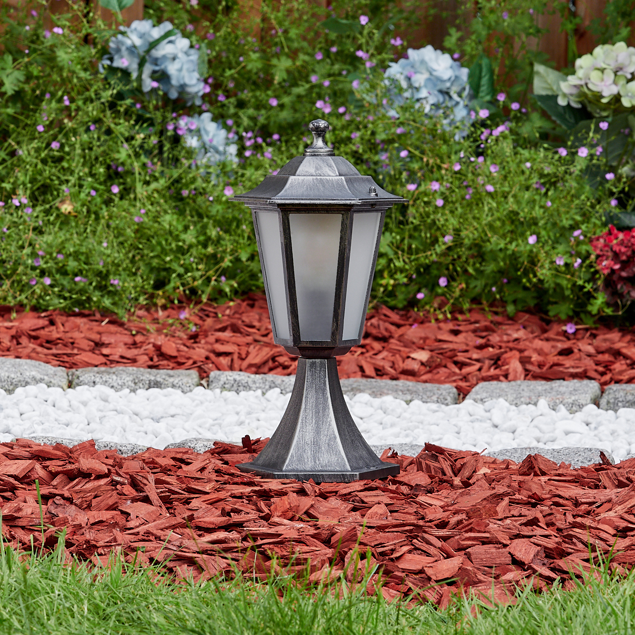 Eine silberne Außenlampe steht auf rotem Mulch. Vordergrund: grünes Gras. Hintergrund: Garten und Holzzaun.