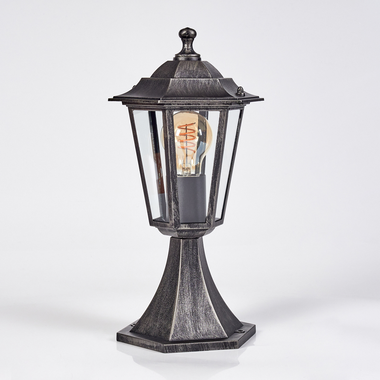 Eine Lampe im antiken Stil mit einem Glaskasten und einer sichtbaren Glühbirne. Die Lampe hat ein metallisches Finish und steht auf einem Sockel.