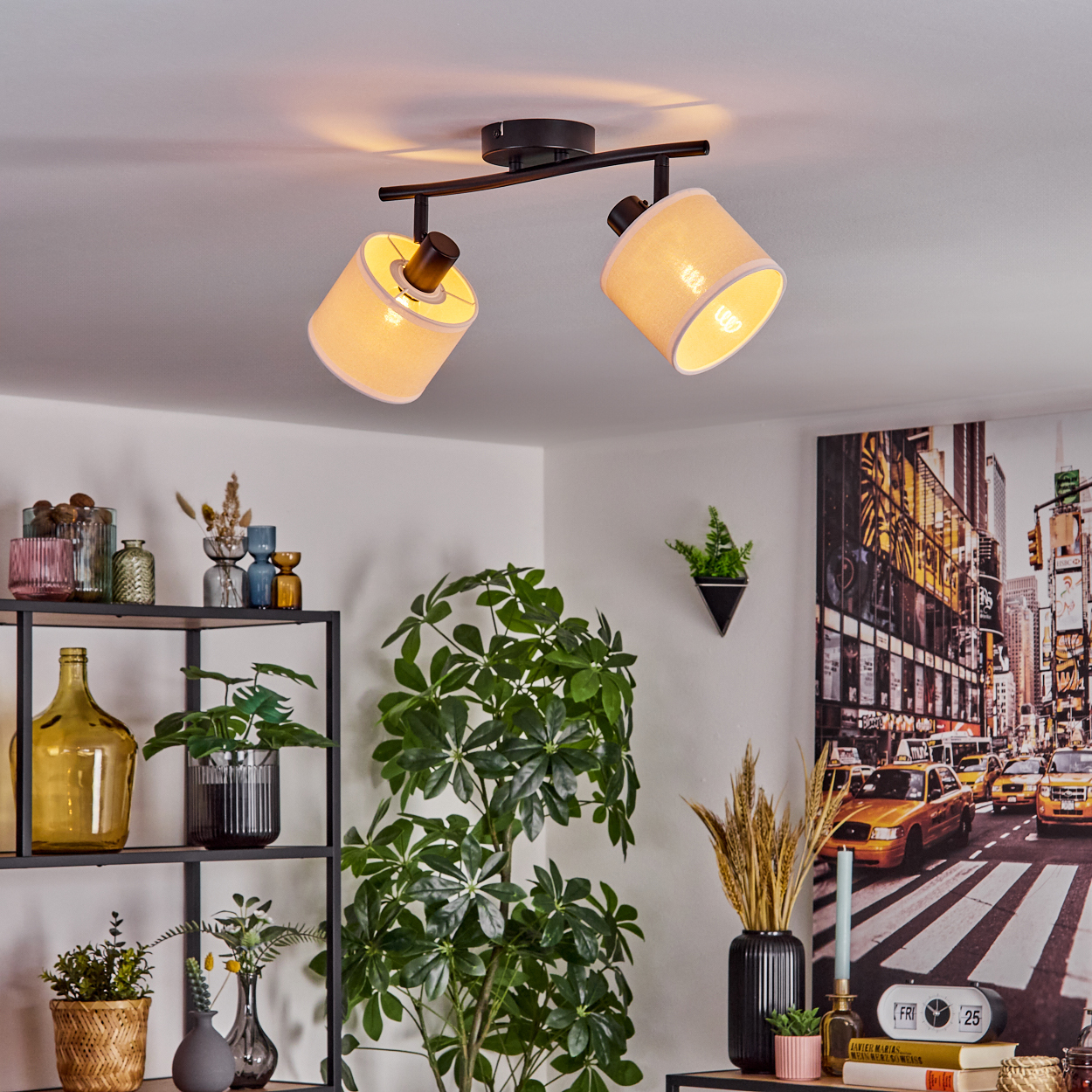 Eine Deckenleuchte mit zwei Lampen beleuchtet ein Zimmer mit Pflanzen, Regalen und einem Stadtbildgemälde. Das Licht ist beige.