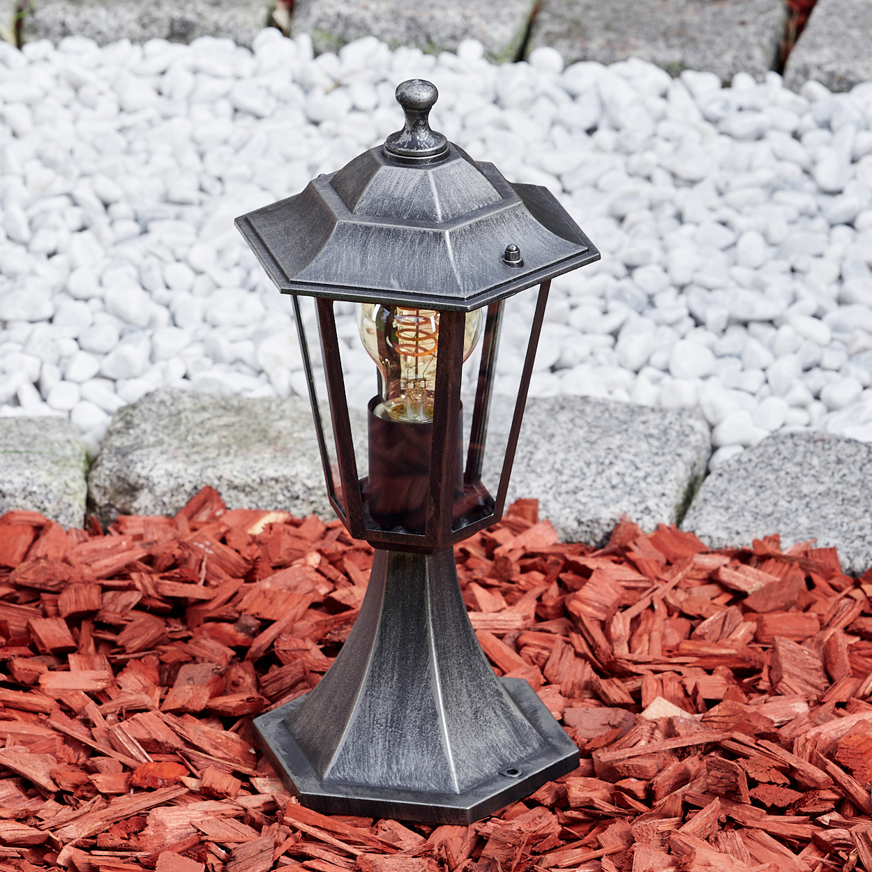 Eine dekorative Außenlampe mit freiliegender Glühbirne steht auf roter Mulch. Im Hintergrund befinden sich weiße Steine und graue Steinblöcke.