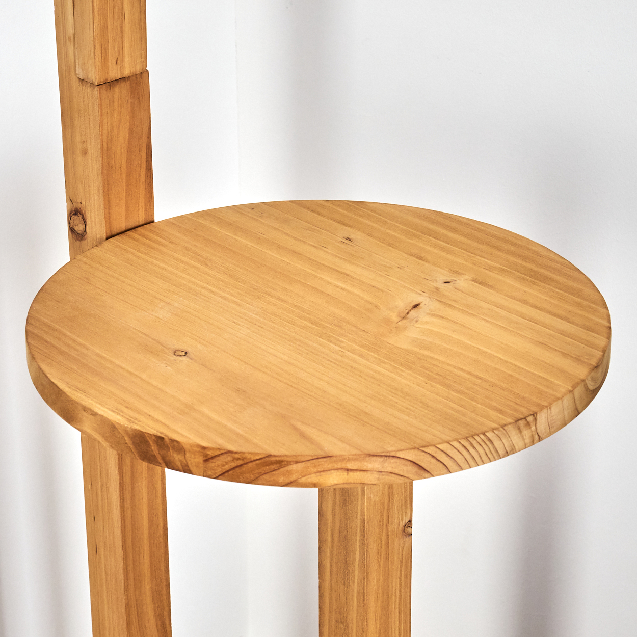 Ein Holzstuhl mit rundem Sitz und rechteckigen Beinen vor einer weißen Wand. Das Holz hat eine hellbraune Farbe.