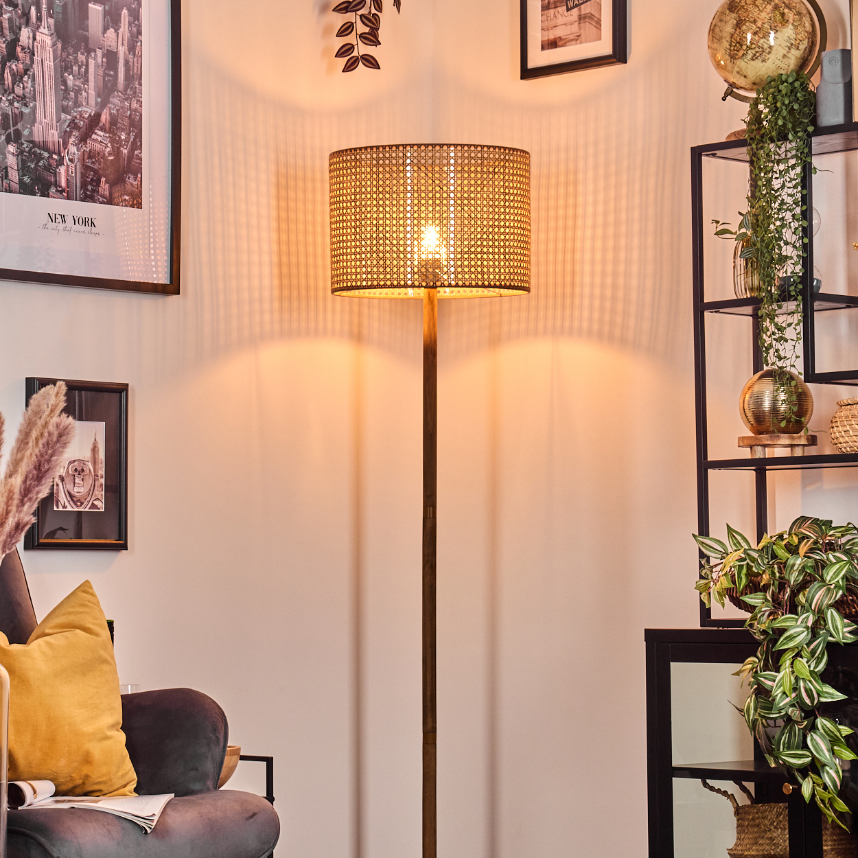 Eine beleuchtete Rattanlampe steht in einem Raum mit Wandkunst, einem Regal mit Pflanzen und einem dunkelgrauen Stuhl mit gelbem Kissen.