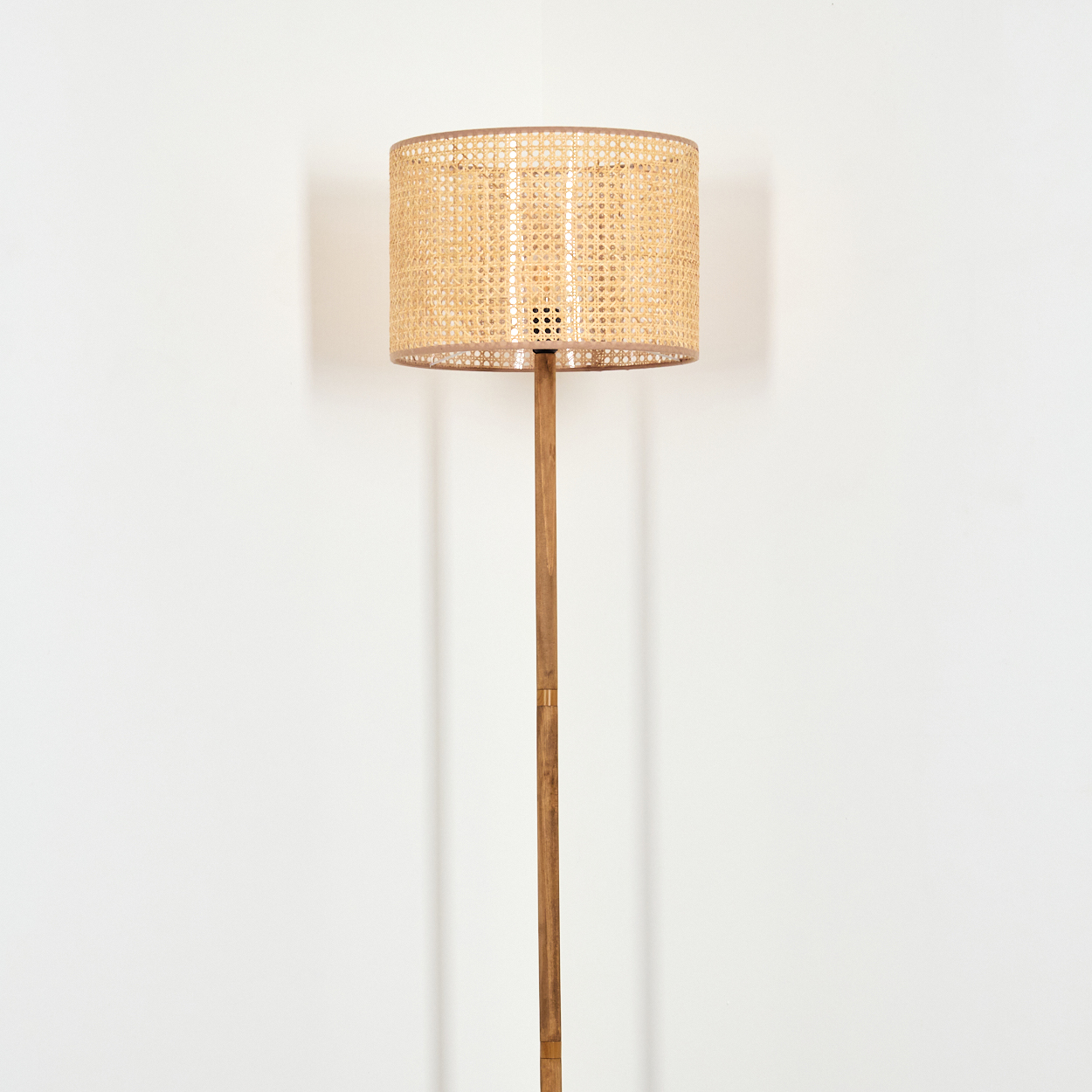 Eine Stehlampe mit gewebtem Lampenschirm und Holzstange steht vor einer weißen Wand. Der Schirm ist zylindrisch und beige.