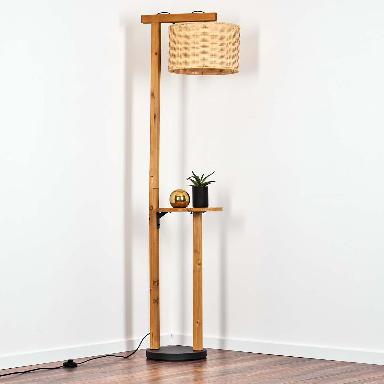 Holz-Stehlampe mit gewebtem Lampenschirm, einem kleinen Regal mit Pflanze und goldener Kugel, vor weißer Wand.